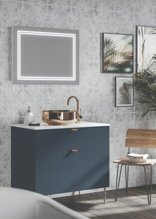 Modern bathroom with decorative tiles, illuminated mirror, and a gold sink. Wooden chair holds towels; green plant adds a natural touch.
