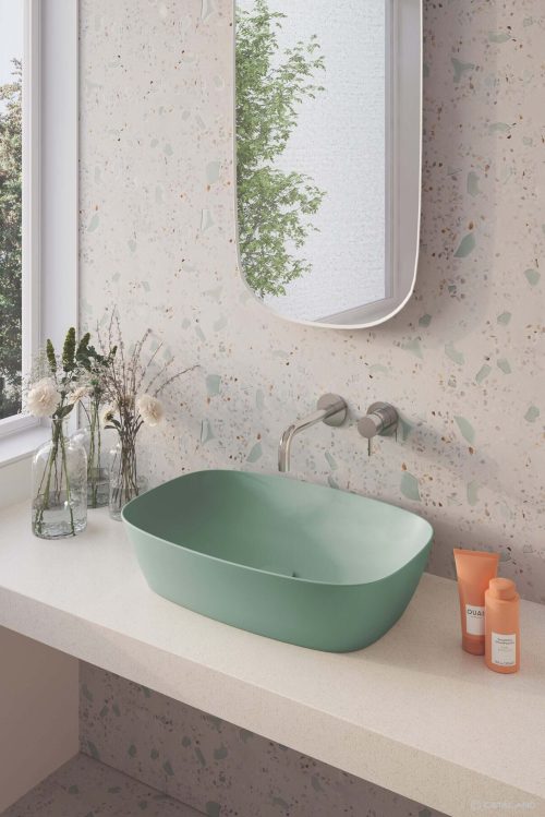 A modern bathroom features a green sink on a terrazzo counter, a wall mirror reflecting greenery, and minimalist decoration with small plants and bottles.