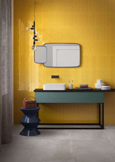 Modern bathroom with yellow tiled wall, abstract mirror, blue vanity, and hanging light fixture. Decorative vase and stacks of plates.
