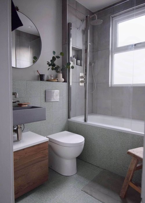 Modern bathroom with gray tiles, a bathtub-shower combo, round mirror, wooden cabinet, toilet, small plants, and natural light from a frosted window.