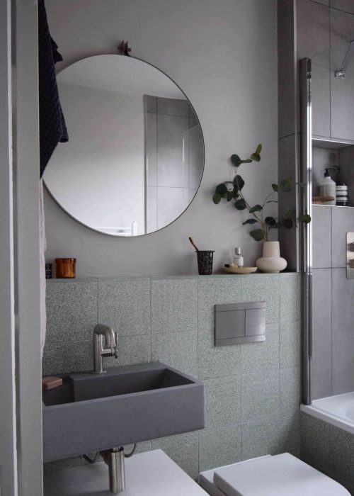 Modern bathroom with a round mirror, gray sink, plants, beige tiles, and a bathtub. Minimalist design featuring subtle decor and neutral colors.