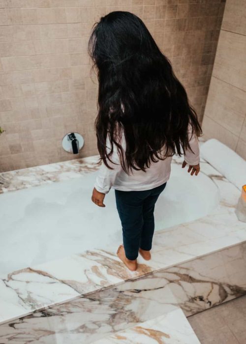 Child standing in a luxurious marble bathtub in a modern bathroom.