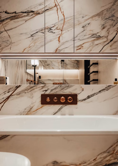 Luxurious marble effect bathroom with warm tones, featuring a bathtub, sleek gold fixtures, and a spacious mirror reflecting ambient lighting, creating an elegant ambiance.