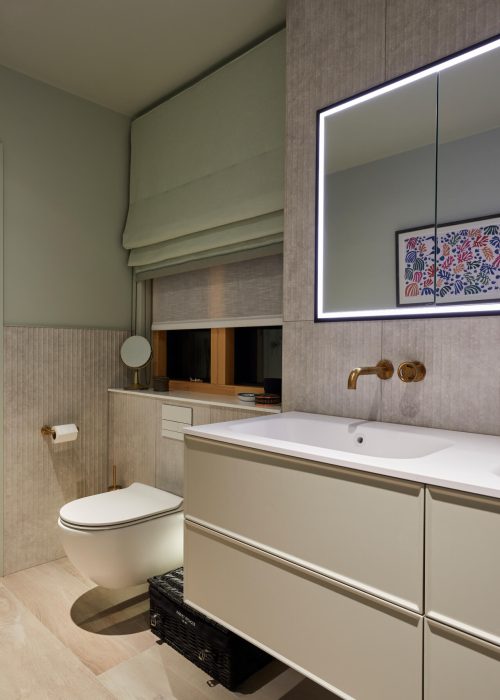 Modern bathroom with warm lighting features a wall-mounted toilet, a sleek vanity, an illuminated mirror, gold fixtures, and a textured wall.