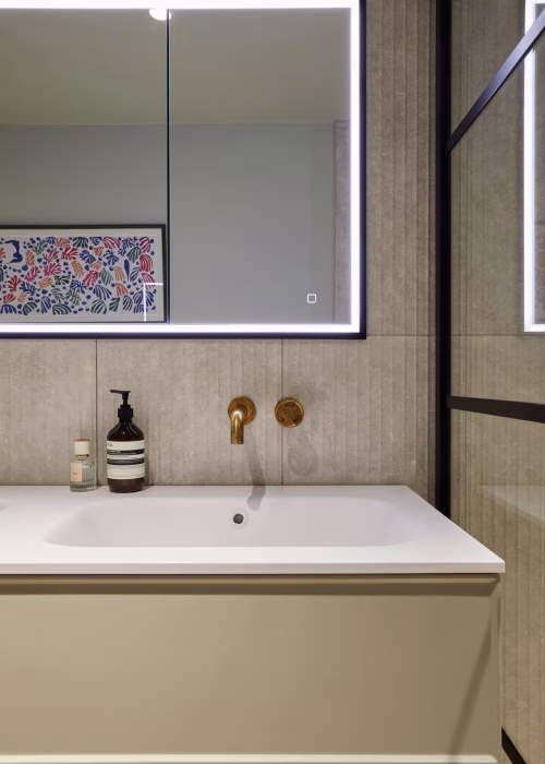 Modern bathroom with a backlit mirror above a white vanity. Gold faucet and soap dispensers are on the counter. Artwork with colorful handprints is reflected.