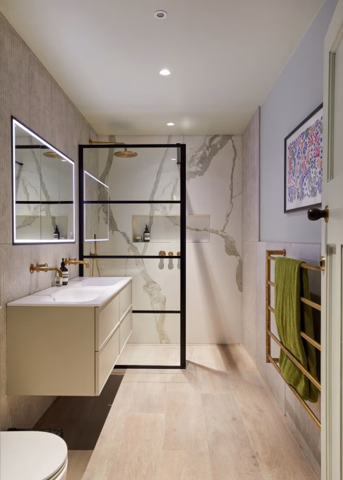 Modern bathroom with a floating vanity and illuminated mirror on the left, glass shower with black frame, light wood flooring, and towel rack on the right.