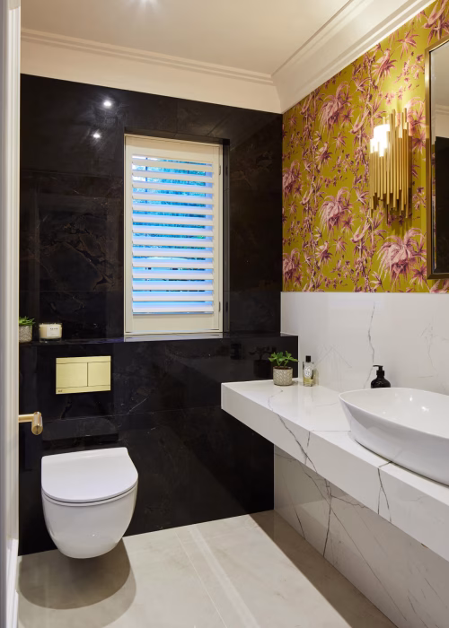 Modern bathroom with a sleek white basin on a marble countertop, floral wallpaper, black marble walls, wall-mounted toilet, and soft lighting.
