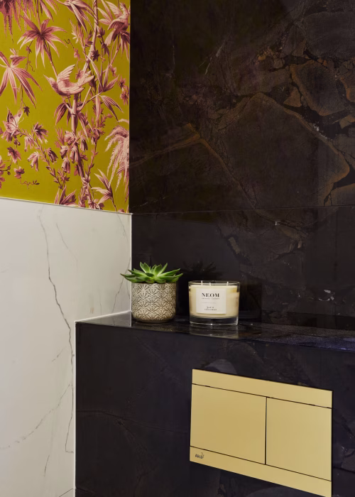 A modern bathroom with dark marble panels and vibrant floral wallpaper. A small potted succulent and a scented candle sit on a shelf, exuding elegance.