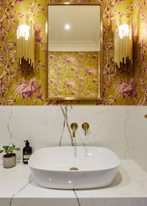 Elegant bathroom with floral wallpaper, a rectangular mirror, and gold sconces. White marble sink, gold faucet, and decorative soap and plant. Warm tone.