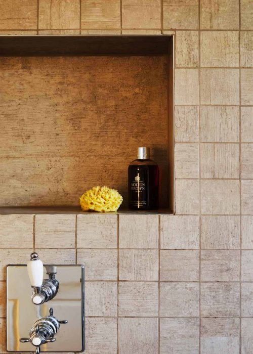 Close-up of a bathroom wall with beige tiles and a built-in shelf holding a black bottle and yellow.