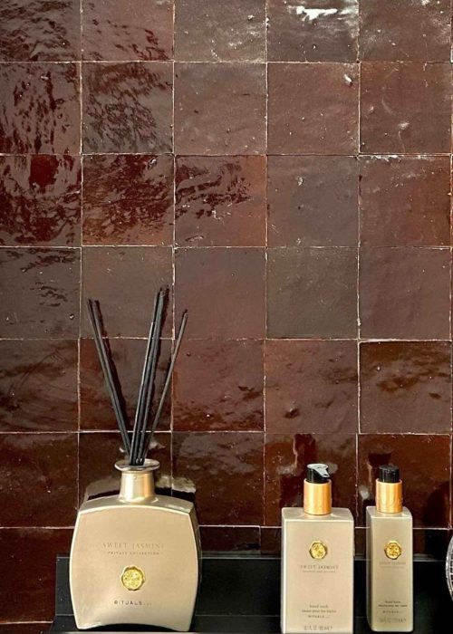 Close-up of glossy brown bathroom tiles with luxury toiletries on a black shelf.