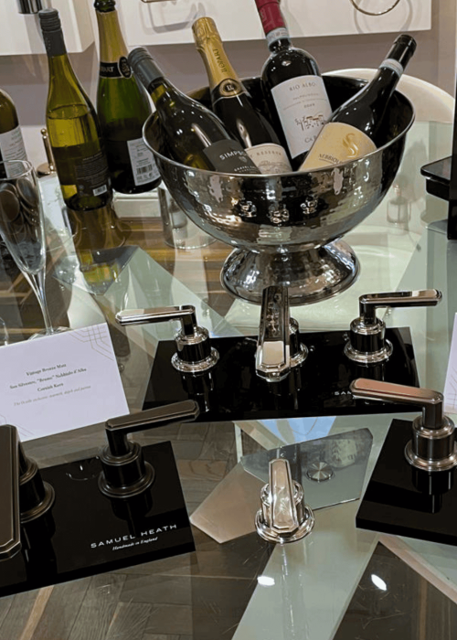 Collection of wine bottles in a silver bowl on a glass bathroom countertop with taps.