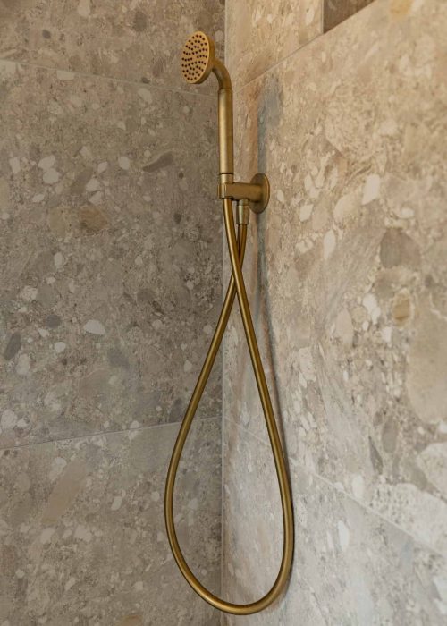 Brass handheld shower head with flexible hose in a stone-tiled bathroom.