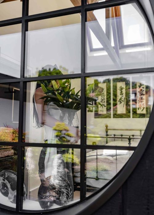 Reflection of a woman holding a plant seen through a decorative window with grid pattern.